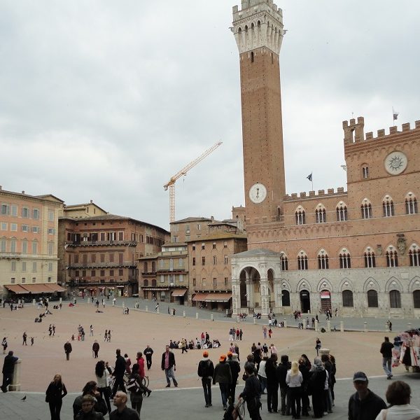 A group of people walking in front of a large building with Piazza del Campo in the background - Tower of Mangia